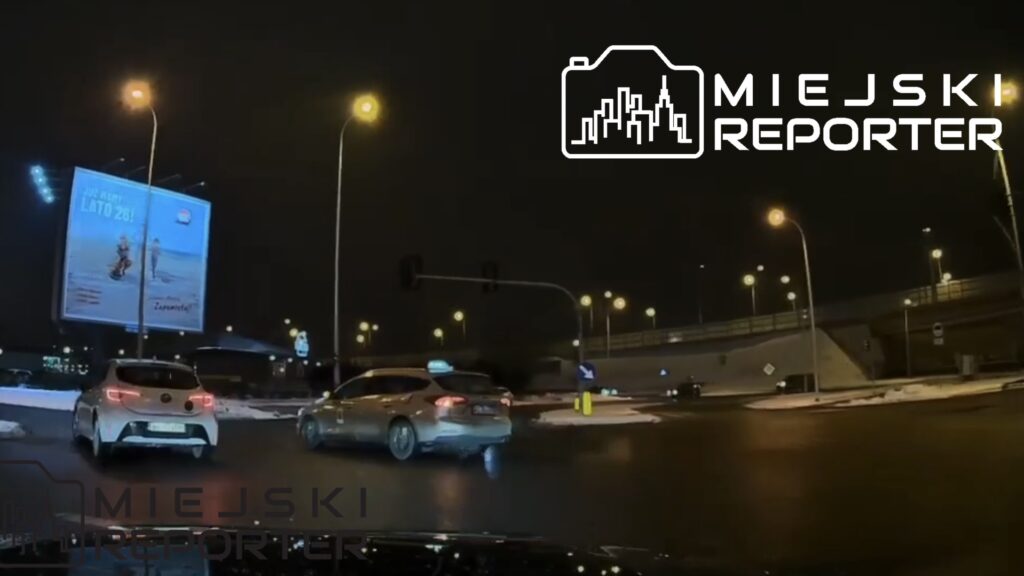 Dwa samochody poruszające się po rondzie w nocy, z oświetlonymi ulicami i reklamą w tle.