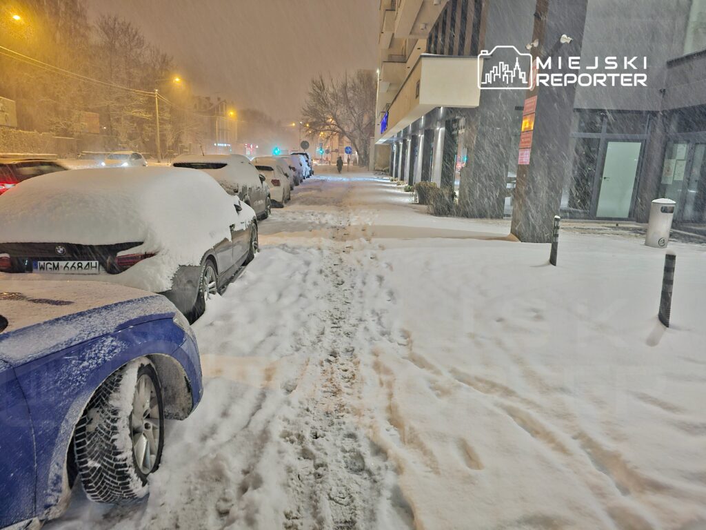 Zasypana śniegiem ulica z zaparkowanymi samochodami, na chodniku widoczne ślady stóp w świeżym puchu.