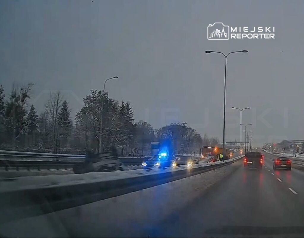 Radiowóz z niebieskimi światłami i pojazd pomocy drogowej na poboczu zaśnieżonej drogi, obok przewrócone auto.