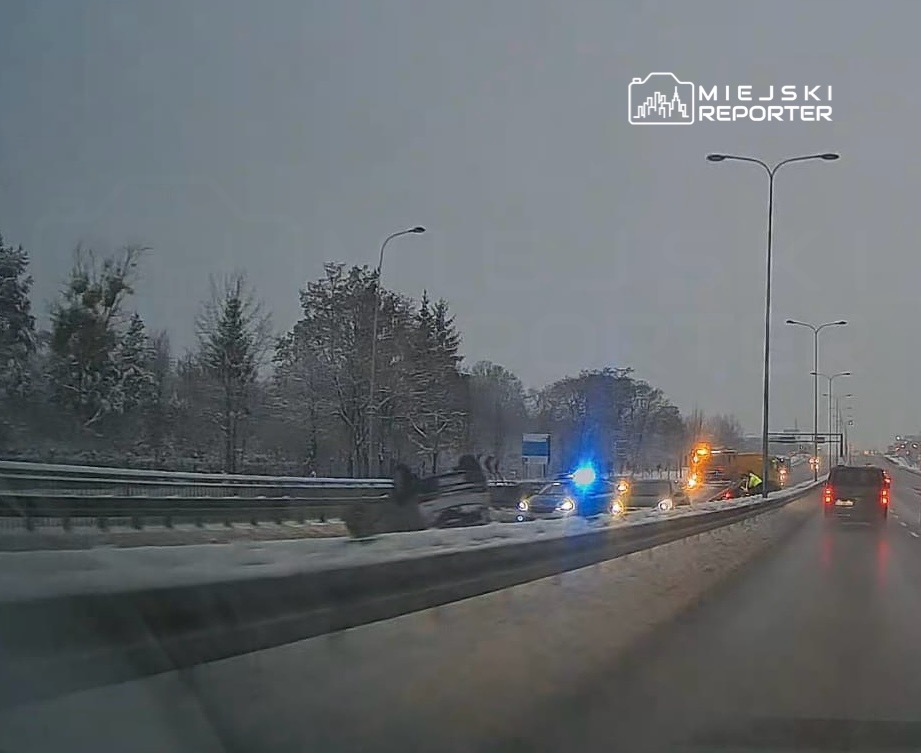 Samochód leży na boku na drodze, obok interweniują radiowozy i pojazdy pomocy drogowej w zimowym otoczeniu.