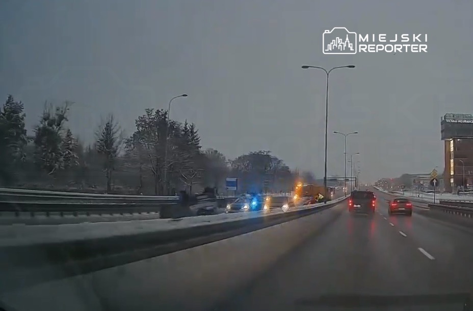 Na zaśnieżonej drodze widać pojazdy z włączonymi sygnałami świetlnymi oraz samochody osobowe poruszające się w tle.