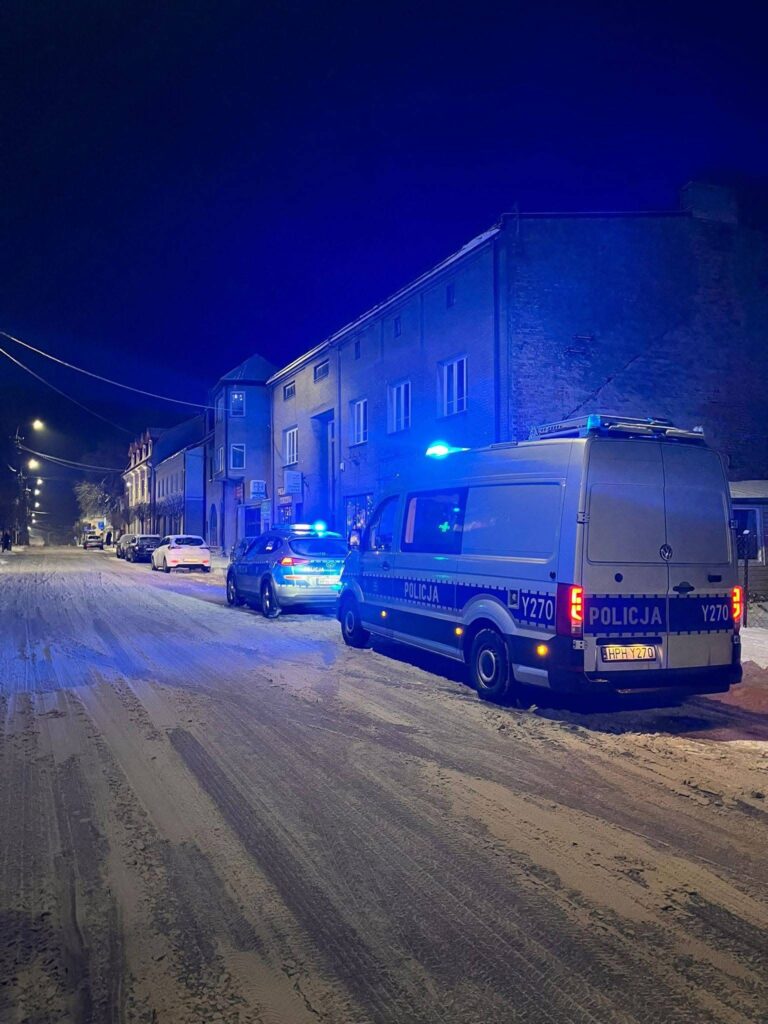 Policja w niebieskich mundurach zabezpiecza ulicę pokrytą śniegiem, obok zaparkowane radiowozy.