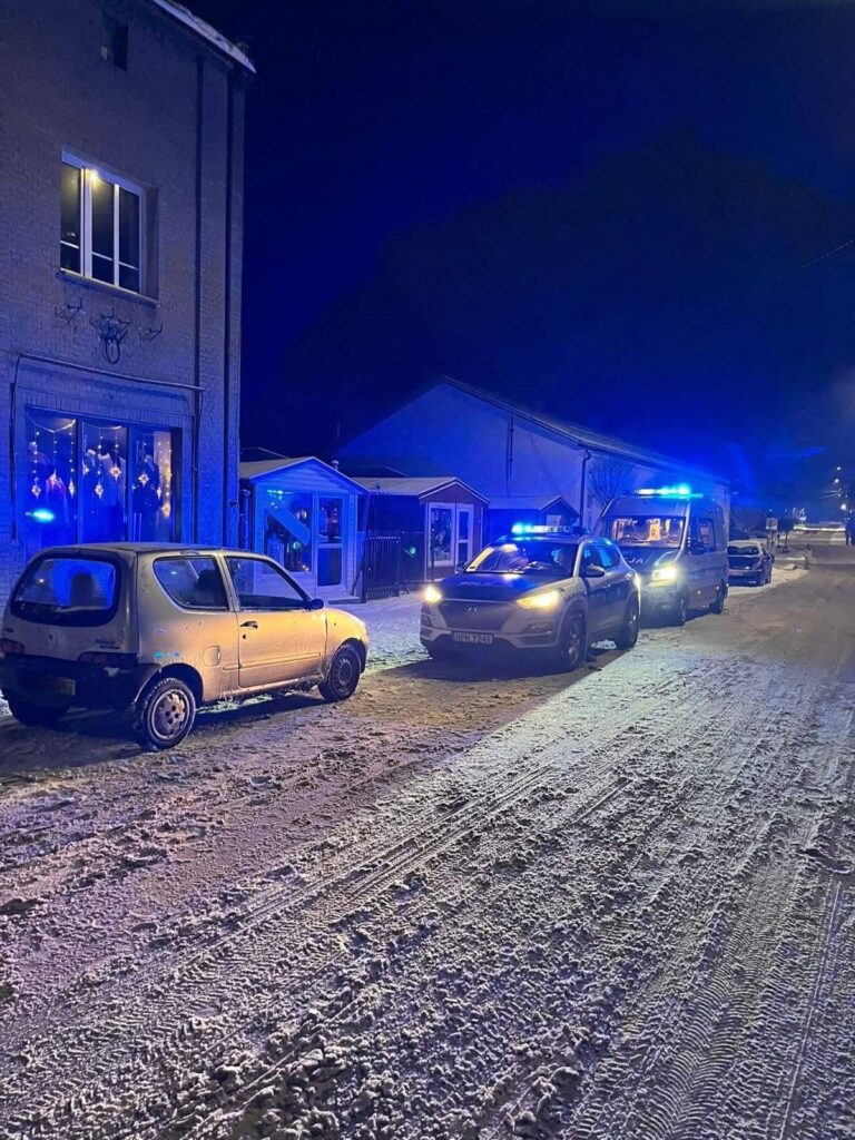 Na oświetlonej nocą ulicy zaparkowane są dwa samochody osobowe oraz radiowóz z włączonymi sygnałami świetlnymi.