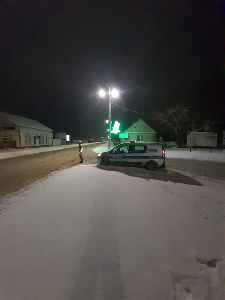 Policjant w odblaskowej kamizelce stoi obok radiowozu na ośnieżonej ulicy w nocy.