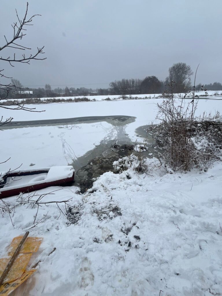 Zamarznięta rzeka z przerwanym lodem, obok leży czerwona łódź, a w tle pokryte śniegiem pola i drzewa.