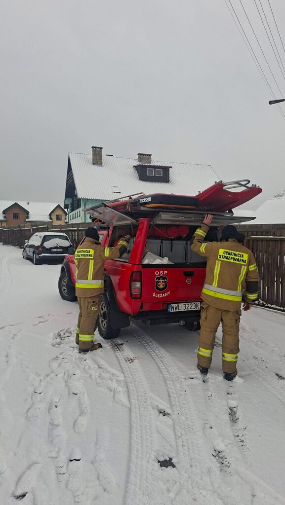 Strażacy w mundurach rozkładają sprzęt z czerwonego wozu strażackiego na zaśnieżonej ulicy.