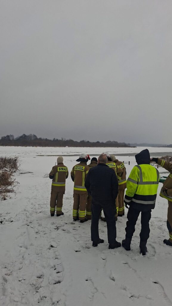 Grupa strażaków w mundurach obserwuje zamarzniętą rzekę pokrytą śniegiem w zimowej scenerii.