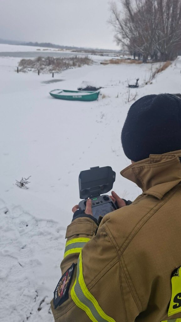 Strażak w zimowym ubraniu obsługuje drona na zaśnieżonym brzegu jeziora, w tle widoczna łódź.