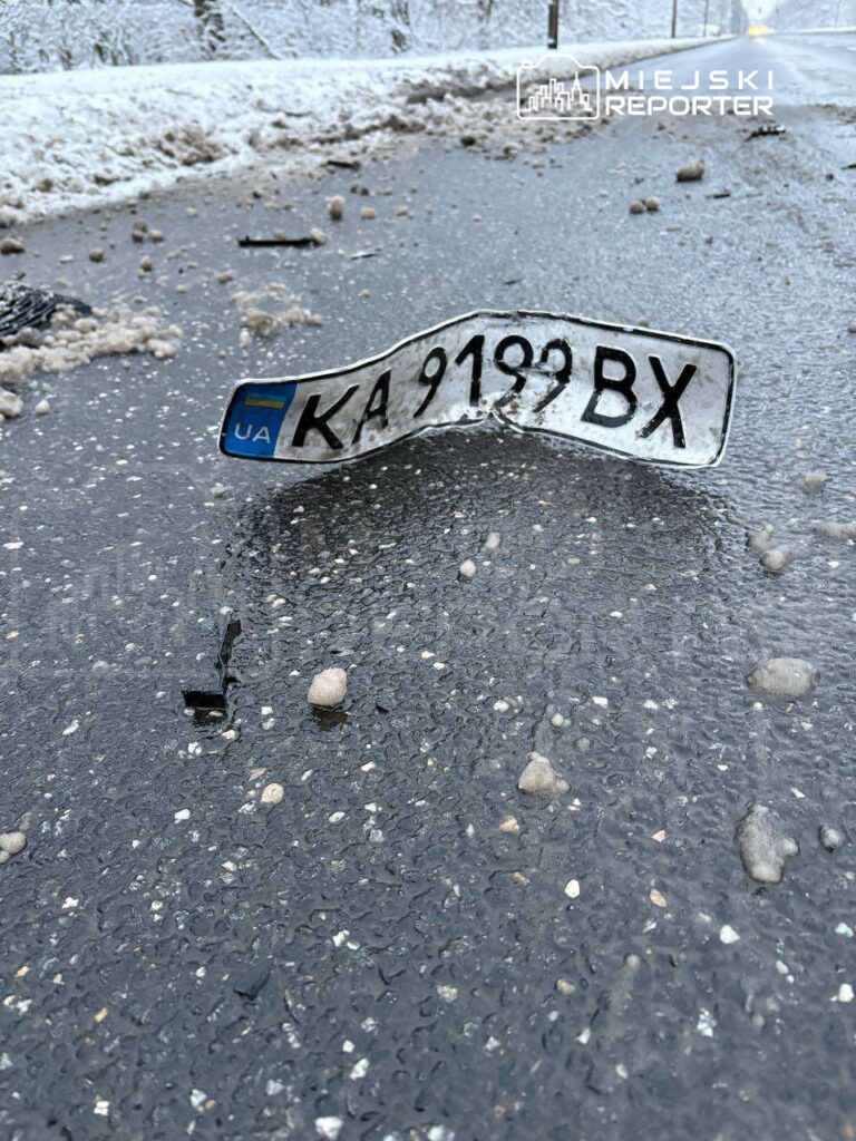 Złamana tablica rejestracyjna leży na zaśnieżonej drodze, otoczona małymi kamieniami i błotem.