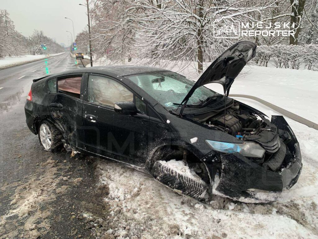 Czarny samochód osobowy z uszkodzeniami stoi na poboczu drogi, z otwartą maską, otoczony śniegiem.