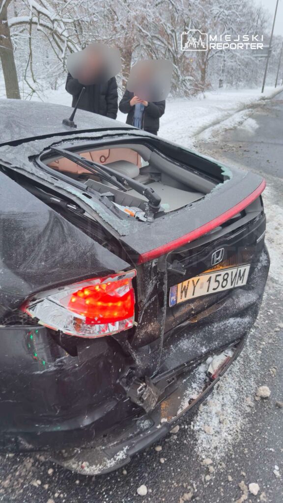 Czarny samochód osobowy z uszkodzoną tylną częścią stoi na zaśnieżonej drodze, obok dwie osoby rozmawiają.