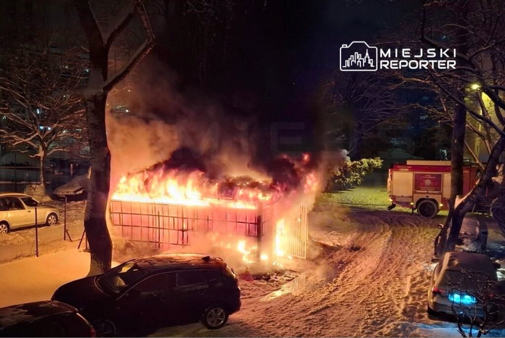 Płonący kontener na śmieci w ośnieżonym otoczeniu, w tle widać woz strażacki oraz zaparkowane samochody.