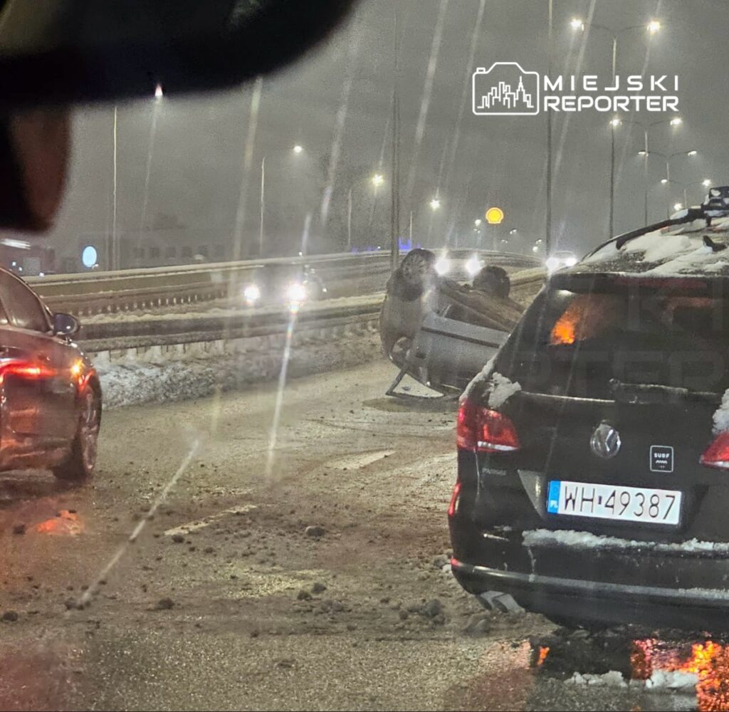 Na drodze leży przewrócone auto, obok zatrzymane pojazdy w deszczowej, zimowej scenerii.