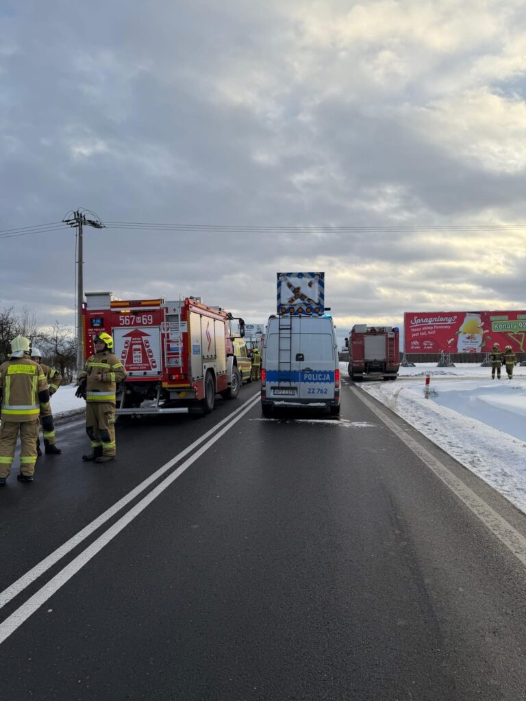 Grupa strażaków i policjantów pracuje przy wypadku drogowym na zaśnieżonej drodze, obok pojazdów ratunkowych.