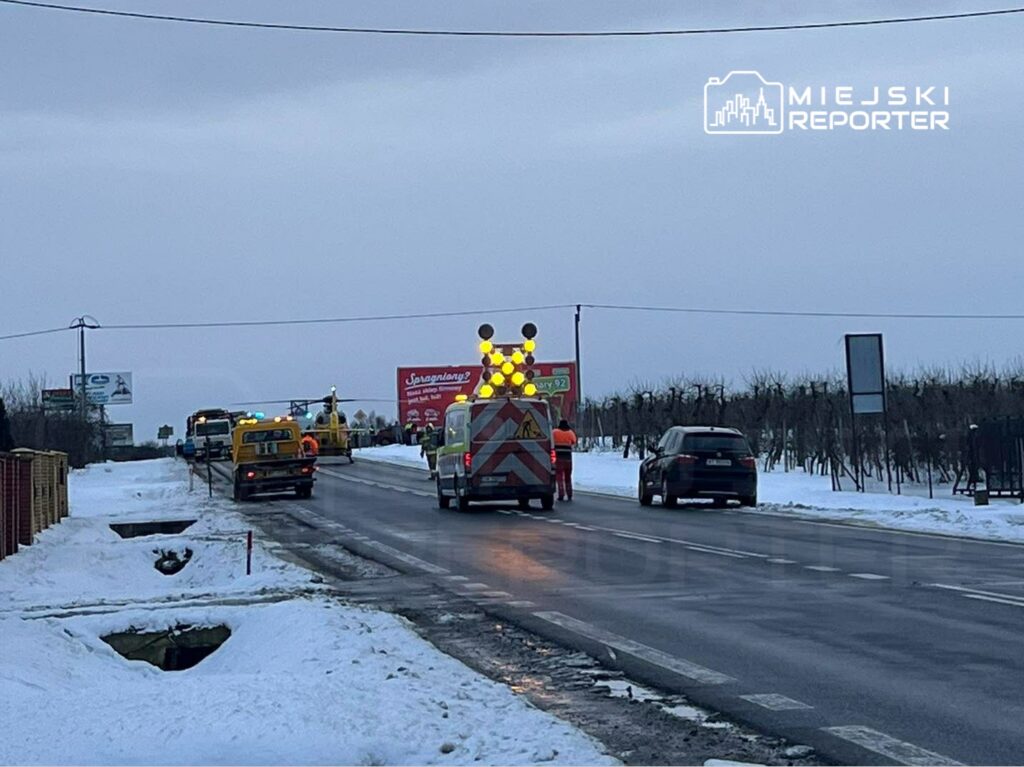 Na drodze w zimowej scenerii pracują pojazdy służb drogowych, a w tle widoczny jest billboard.