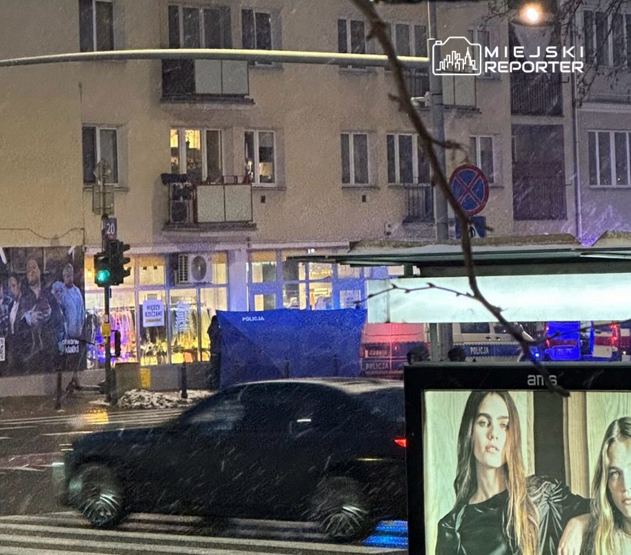 Czarny samochód osobowy przejeżdża obok stoiska miejskiego w trakcie opadów śniegu, w tle widać policyjny namiot.