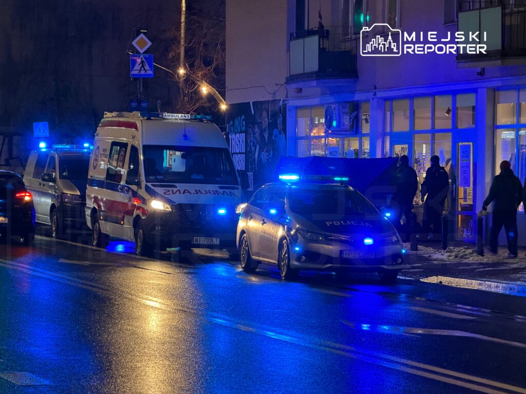 Na ulicy stoją karetka pogotowia i radiowóz policyjny z włączonymi sygnałami świetlnymi, obok grupka ludzi.