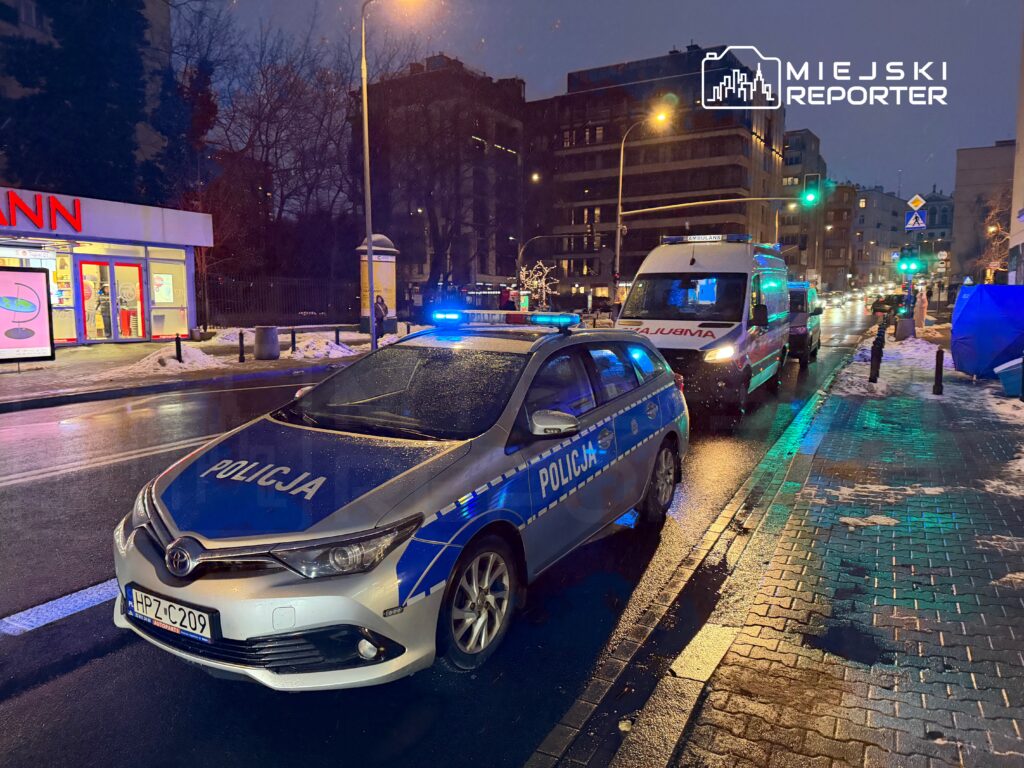 Policyjny samochód z niebieskimi światłami zaparkowany na ulicy, obok ambulans i sklep z reklamą.