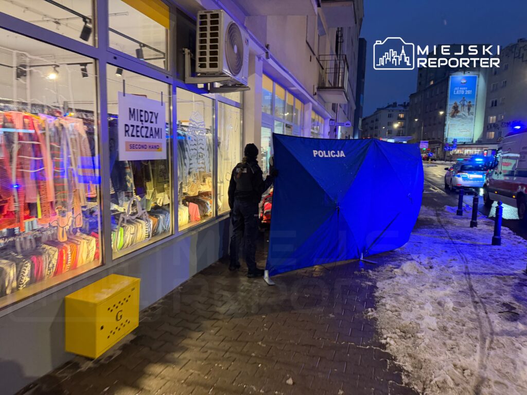 Policjant rozmawia z osobą przy niebieskim namiocie z napisem "POLICJA" na chodniku obok sklepu z odzieżą używaną.