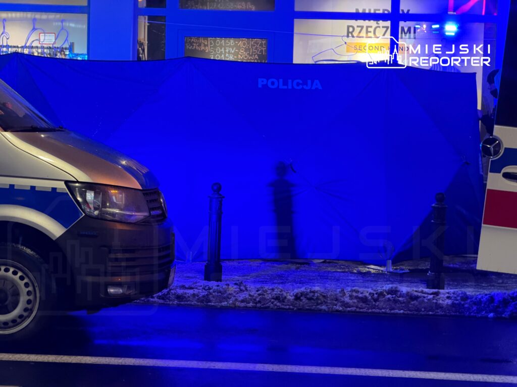 Policyjny van zaparkowany przy niebieskim namiocie, w tle widoczna ulica i zasypana śniegiem latarnia.