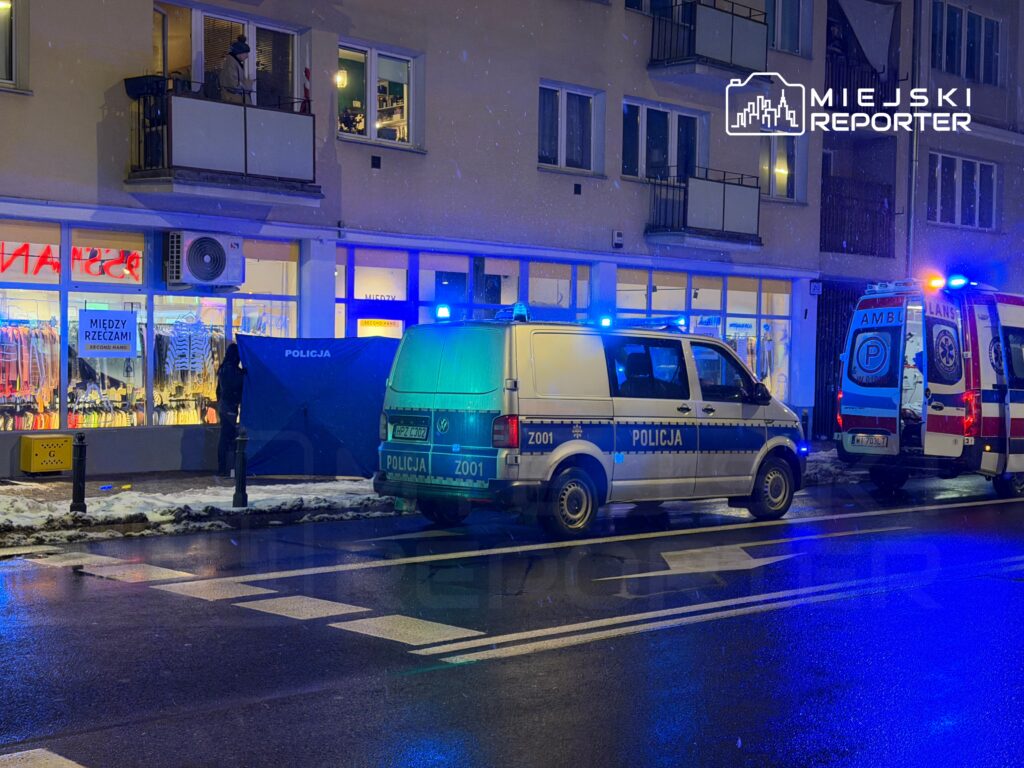 Policja i karetka pogotowia przed sklepem odzieżowym, w tle osoba na balkonie, nocna sceneria z padającym śniegiem.