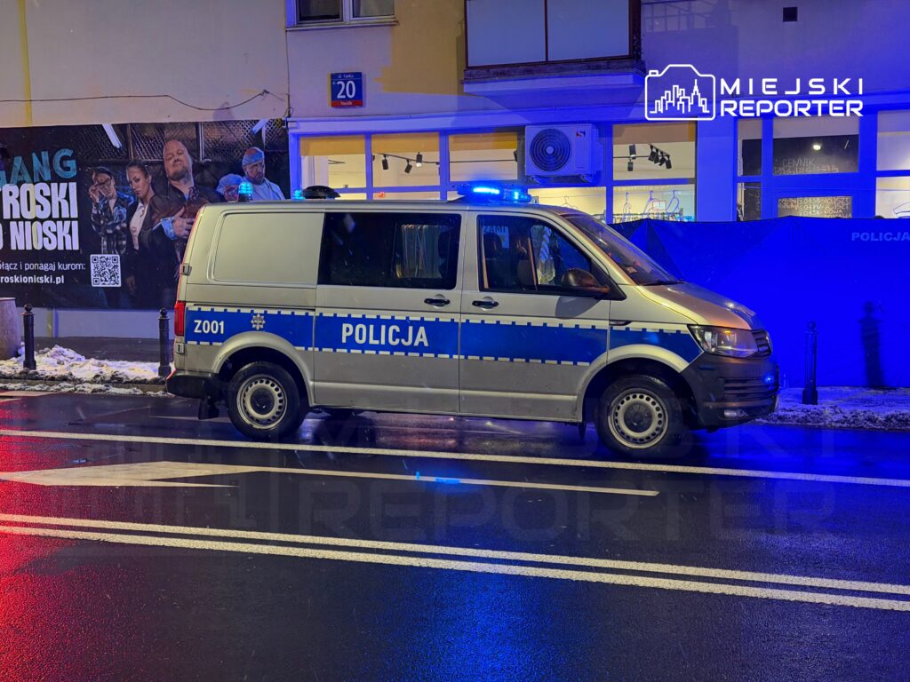 Policyjny van z włączonymi sygnalizatorami świetlnymi stoi na ulicy obok dużego billboardu reklamowego.