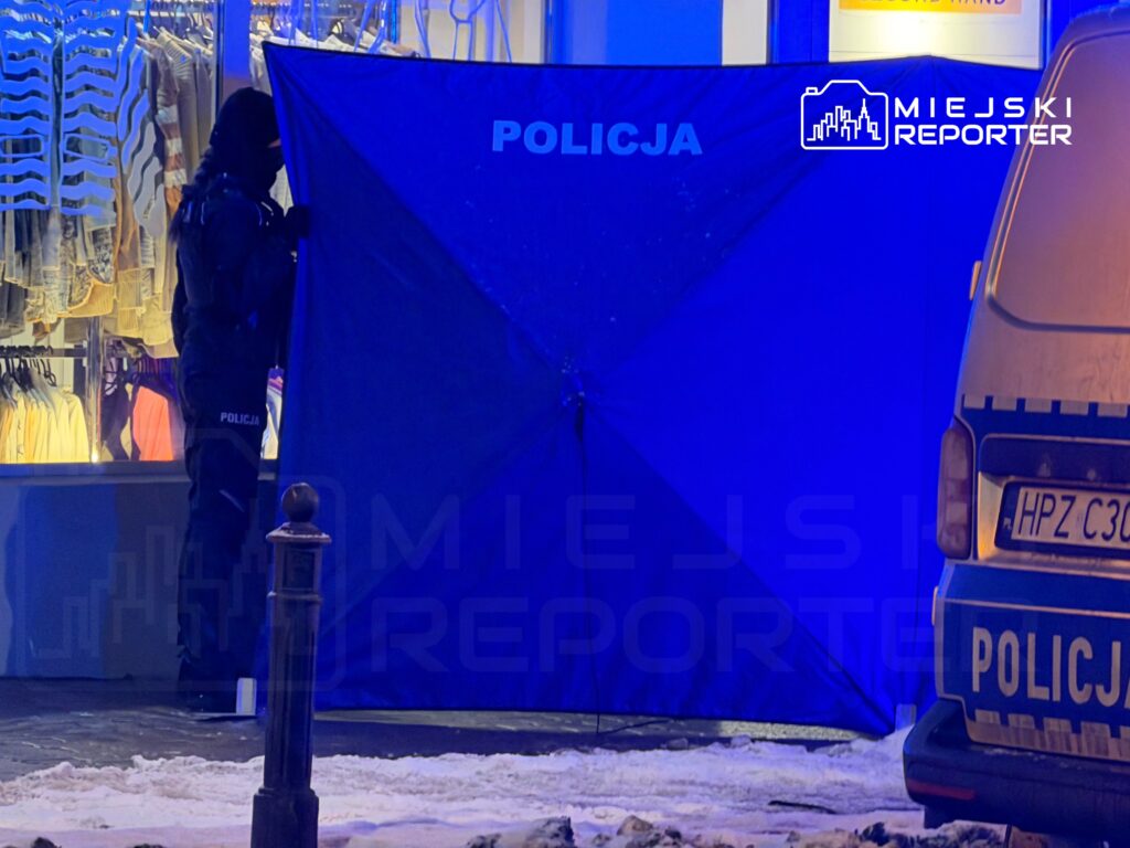 Policjant w czarnym mundurze ustawia niebieską osłonę przed witryną sklepową w zaśnieżonym otoczeniu.