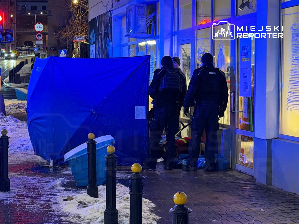 Policjanci w mundurach udzielają pomocy osobie leżącej na chodniku przed sklepem, w tle widoczne oświetlenie uliczne.