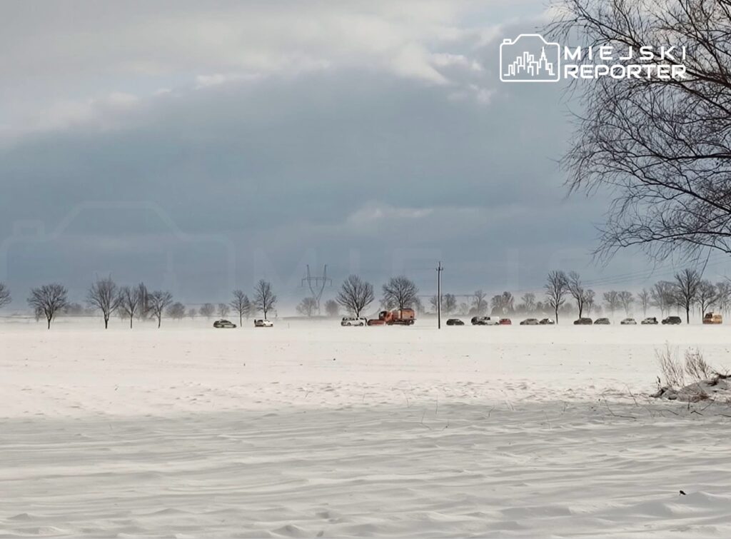 Pojazdy stojące w zaspach śnieżnych na otwartej przestrzeni, w tle drzewa i chmury.