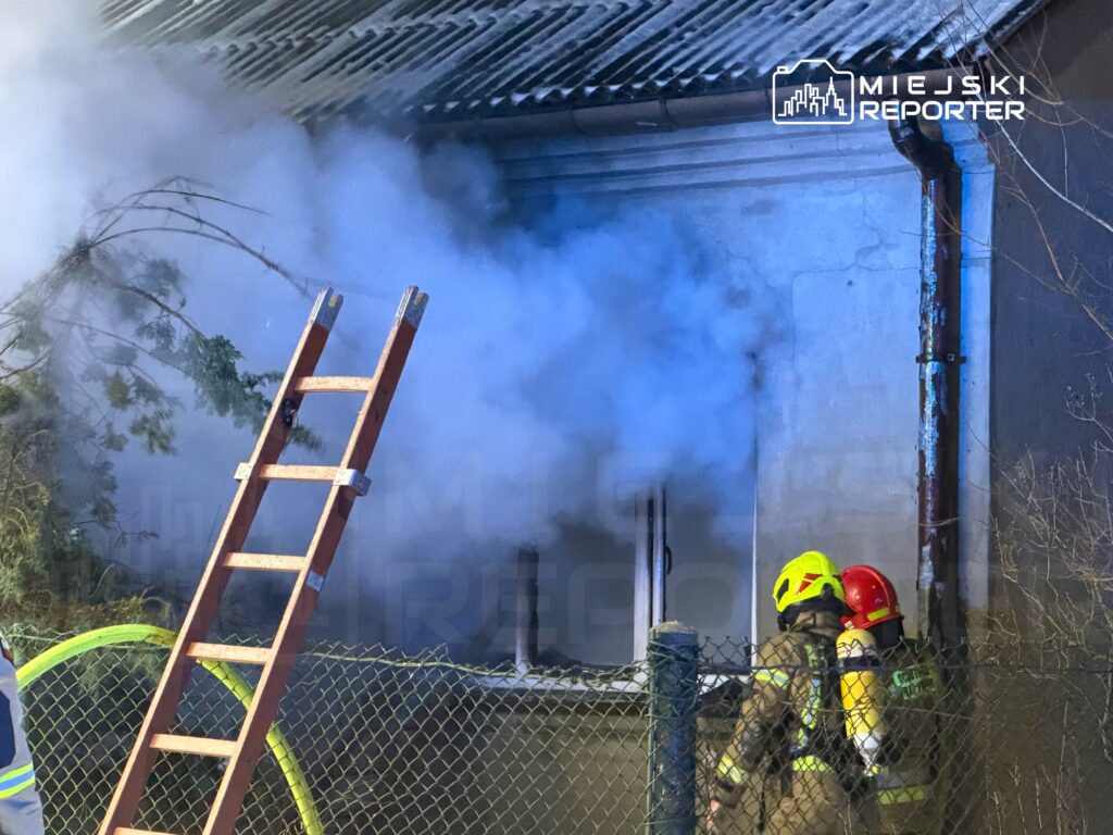 Strażacy w hełmach, jeden w żółtym, drugi w czerwonym, pracują przy zadymionej ścianie budynku, obok drabina.
