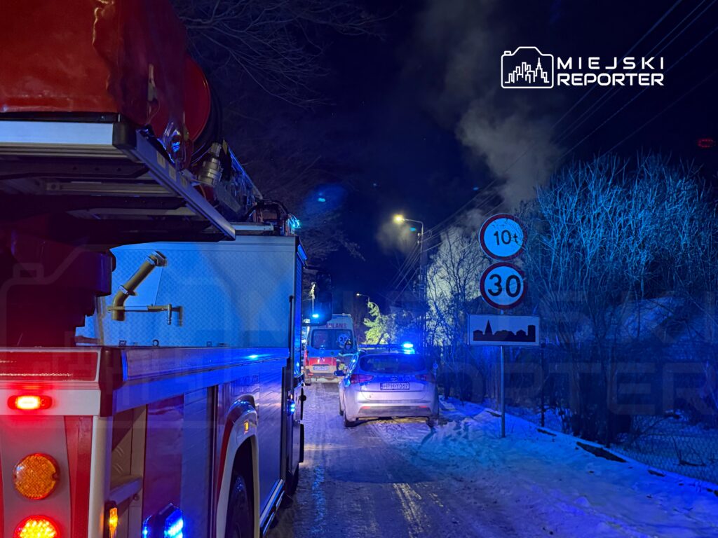 W nocy widać wóz strażacki z włączonymi sygnałami, obok pojazd policyjny oraz unoszący się dym w tle.