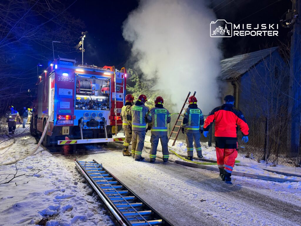 Grupa strażaków w hełmach i odzieży ochronnej pracuje przy pożarze w budynku, obok wozu strażackiego na zaśnieżonej drodze.