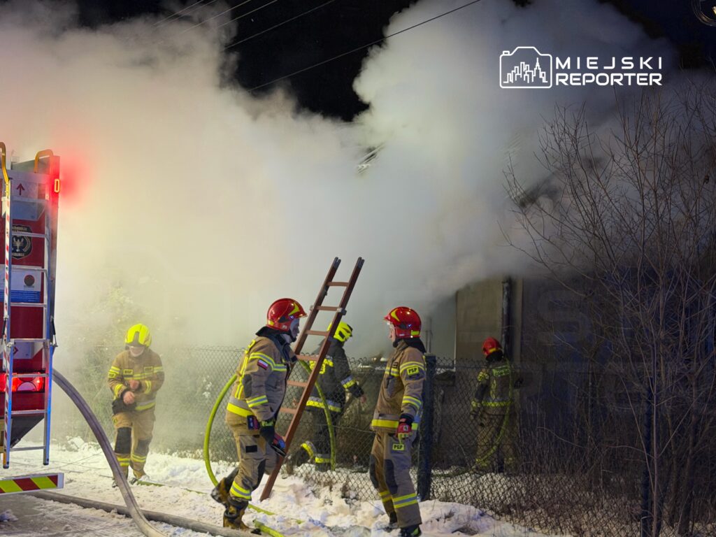 Strażacy w hełmach i mundurach gaszą pożar, otoczeni gęstym dymem i śniegiem, przy ogrodzeniu.