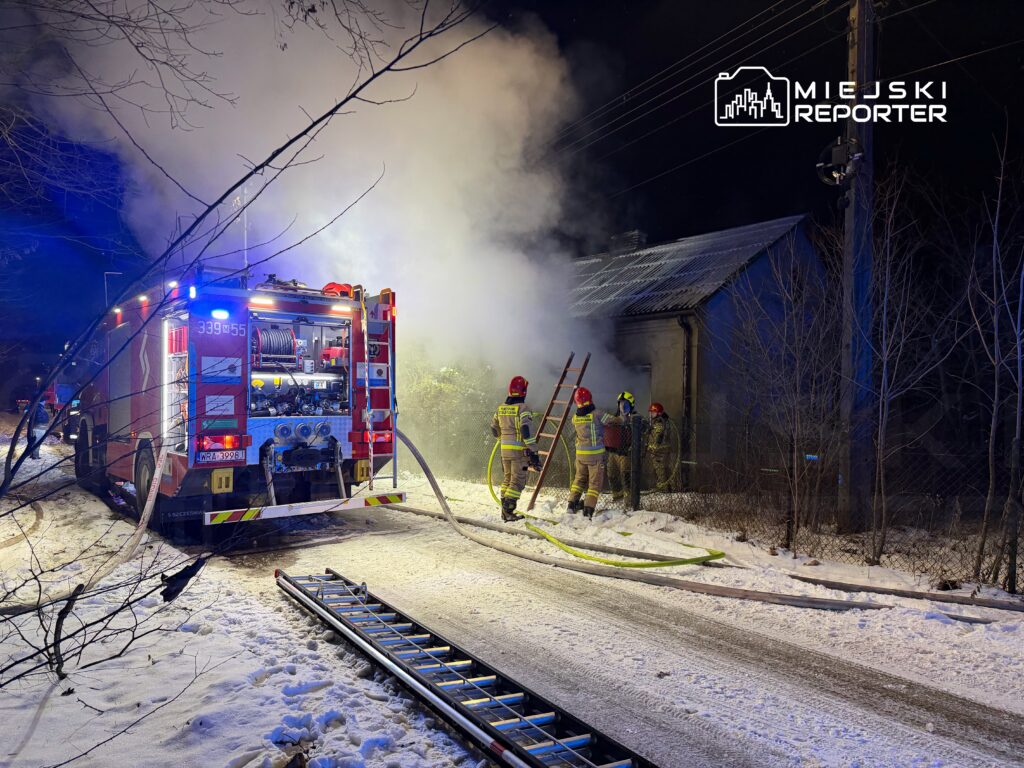 Strażacy w hełmach gaszą pożar w budynku, obok widać wóz strażacki i leżące na ziemi węże.