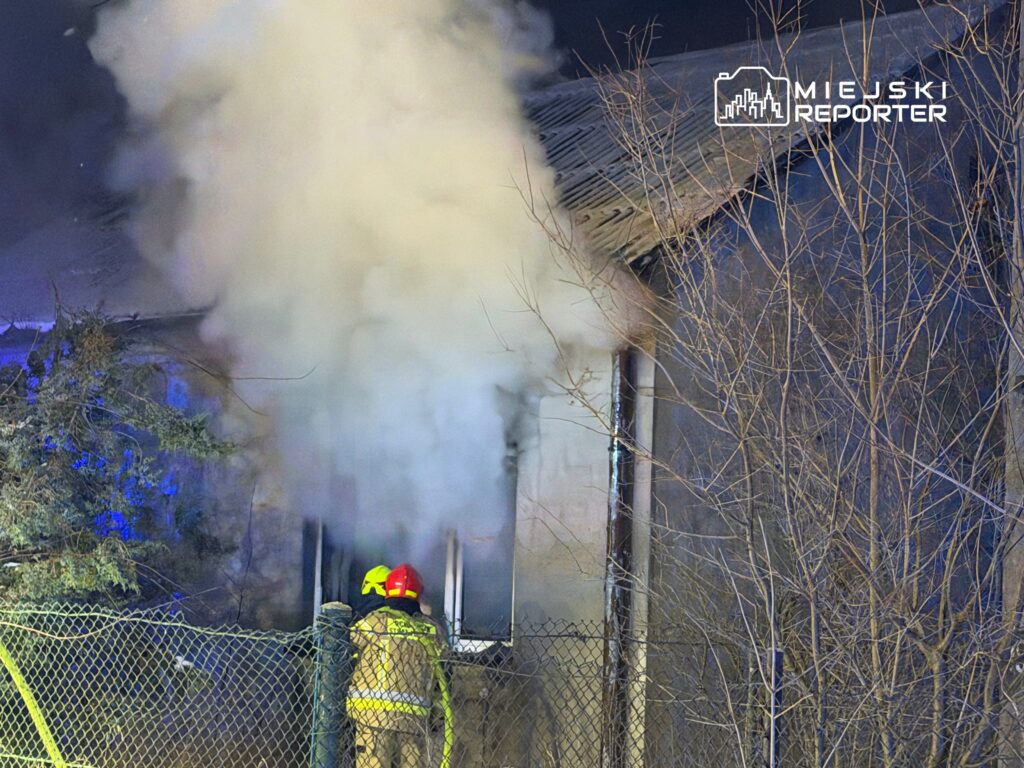 Strażacy w hełmach gaszą pożar w budynku, z którego wydobywa się gęsty dym. W tle widoczne drzewa i ogrodzenie.