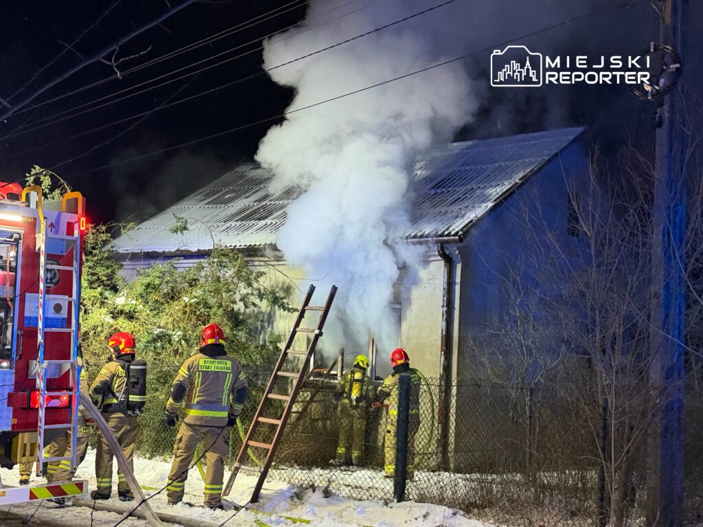 Grupa strażaków w hełmach gasi pożar domu, z którego wydobywa się gęsty dym, w otoczeniu zimowego krajobrazu.