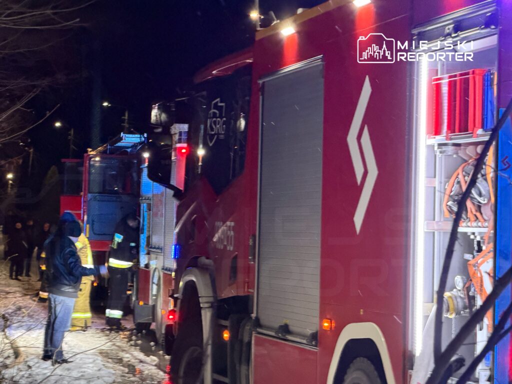 Strażacy w żółtych kombinezonach pracują przy wozie strażackim w nocnej scenerii, otoczeni przez ludzi i drzewa.