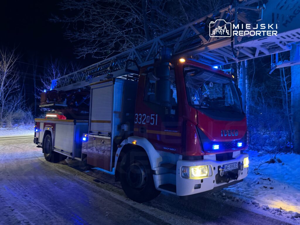 W nocy w lesie stoi wóz strażacki z podnośnikiem, oświetlony niebieskimi światłami.