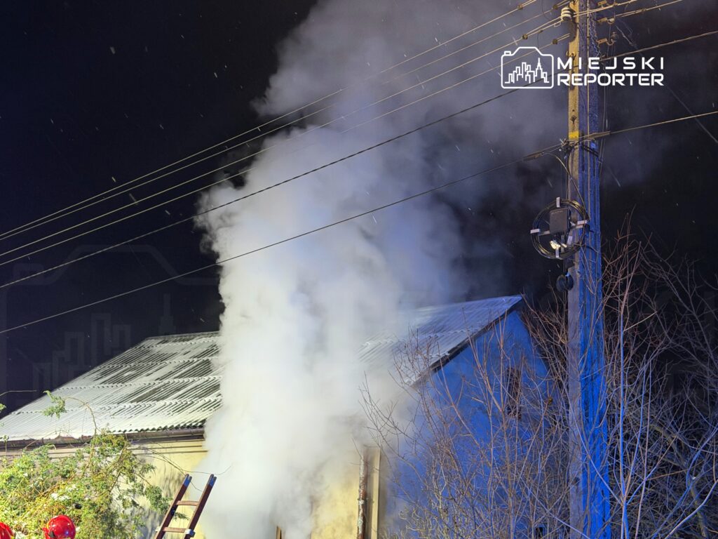 Gęsty, biały dym wydobywający się z budynku w nocy, w otoczeniu linii energetycznych i drzew.