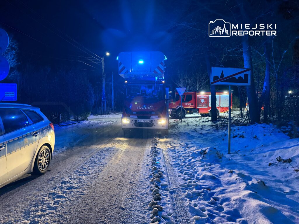 Na ośnieżonej drodze stoi wóz strażacki z włączonymi sygnałami świetlnymi, obok zaparkowany radiowóz policyjny. W tle widoczne są drzewa i znaki drogowe.