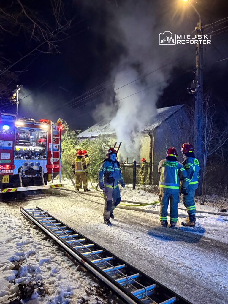 Strażacy w mundurach gaszą pożar w nocy przy zniszczonym budynku, otoczeni przez dym i śnieg.