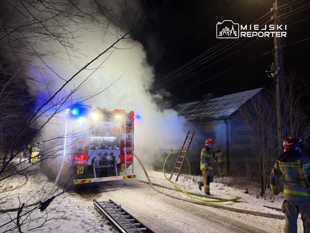 Strażacy w hełmach gaszą pożar przy budynku, obok stoi wóz strażacki z włączonymi sygnałami świetlnymi.