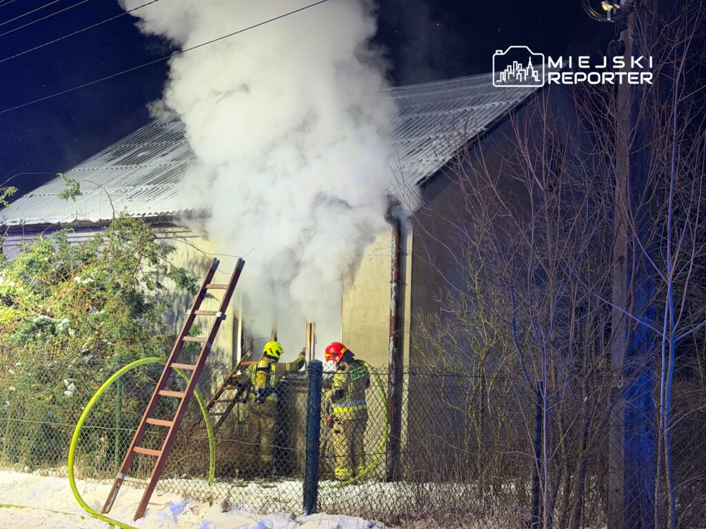 Dwaj strażacy w hełmach gaszą pożar w budynku, z którego wydobywa się gęsty dym, otoczeni przez śnieg i drzewa.
