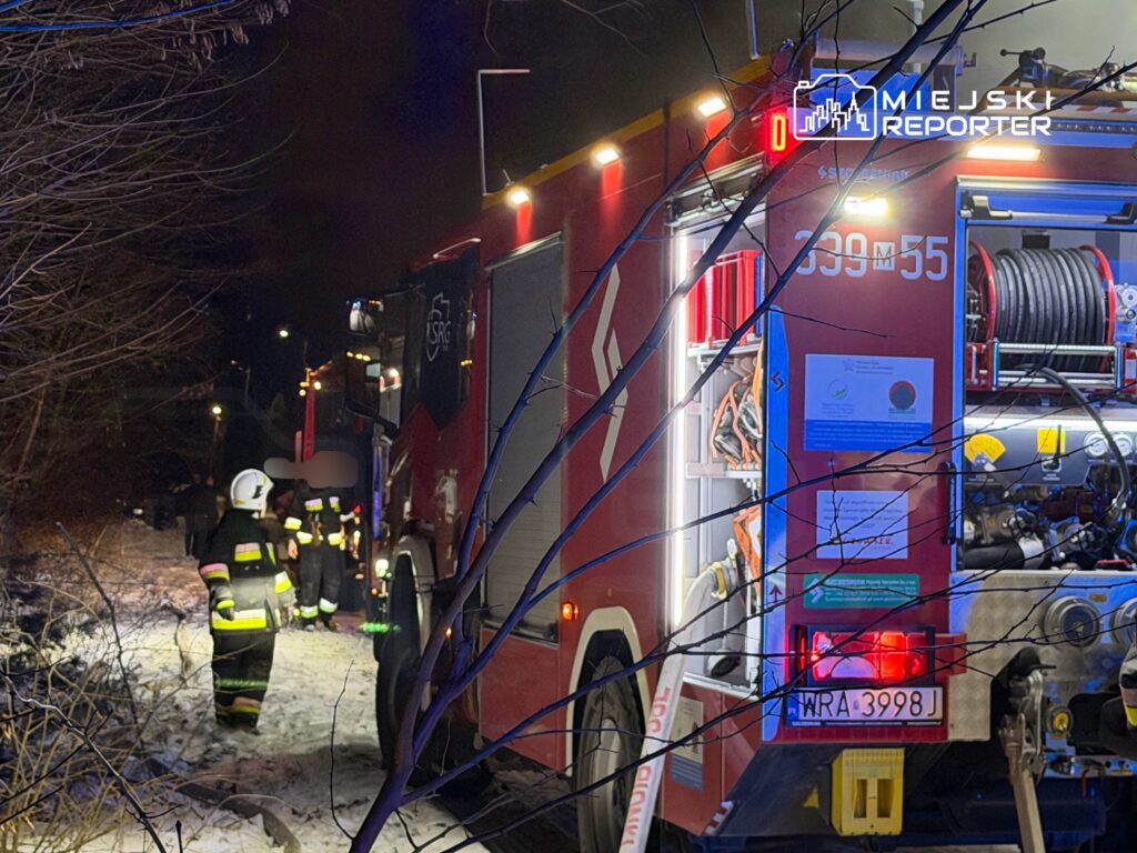 Strażacy w mundurach podchodzą do wozu strażackiego na ośnieżonej drodze w nocy, otoczeni przez drzewa.