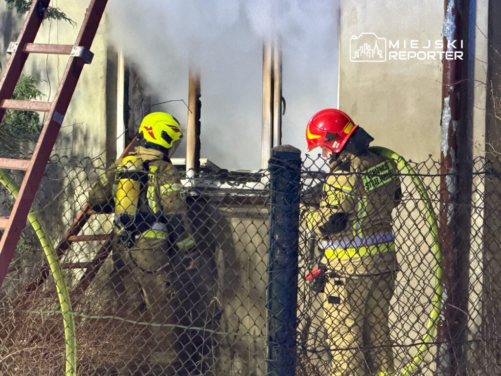 Dwaj strażacy w hełmach gaszą pożar w budynku, otoczonym przez siatkę ogrodzeniową.