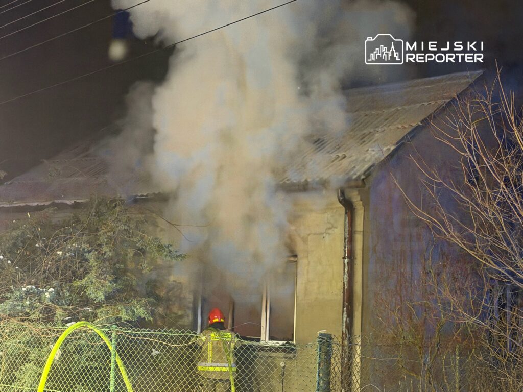 Strażak w hełmie obserwujący dym wydobywający się z okna budynku mieszkalnego w nocy.