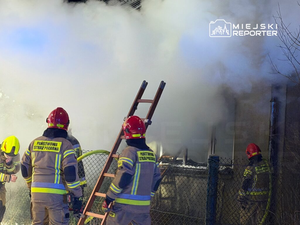 Strażacy w hełmach gaszą pożar w zadymionym otoczeniu, obok drabiny i ogrodzenia.