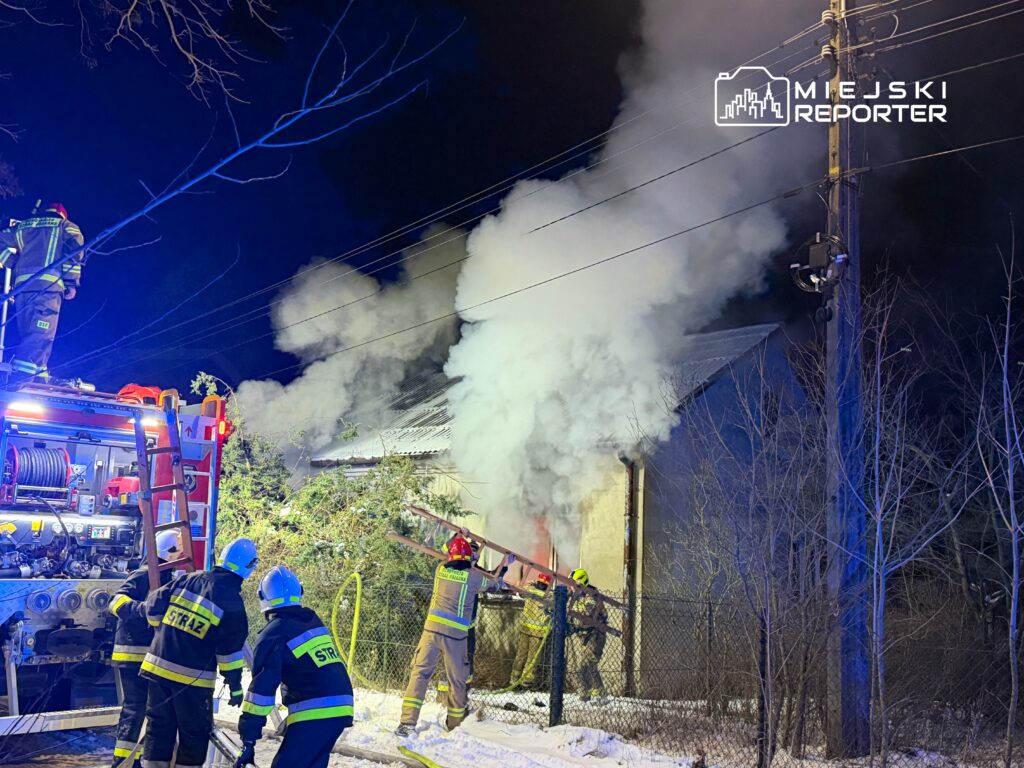 Grupa strażaków gasząca pożar budynku w otoczeniu dymu, w nocy, przy użyciu węża strażackiego.