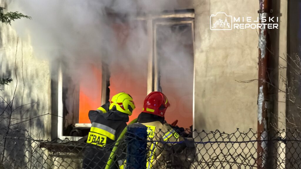 Dwaj strażacy w kaskach gaszą pożar w oknie budynku, otoczeni gęstym dymem.
