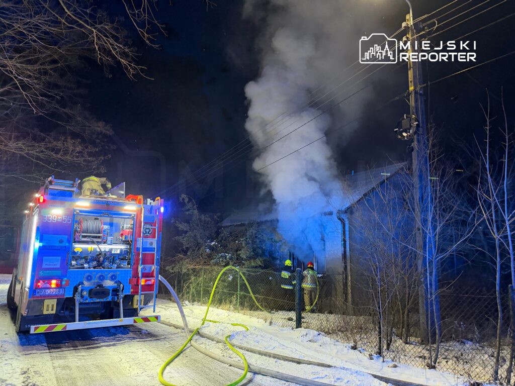 Wóz strażacki z włączonymi sygnałami świetlnymi stoi na ośnieżonej drodze, a strażacy gaszą pożar wydobywający się z budynku.